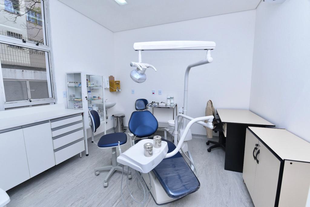 FOTO DE CLÍNICA - uma cadeira odontológica rodeada de equipamentos ocupa o centro da sala com paredes brancas e mobiliário claro, do lado esquerdo temos uma janela com vista para a rua onde há um prédio residencial e árvores